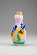 A small hand-blown glass perfume bottle with a cork stopper, decorated with colorful swirls of blue, yellow, green, red, and white on a pale pink base, set against a dark textured background.