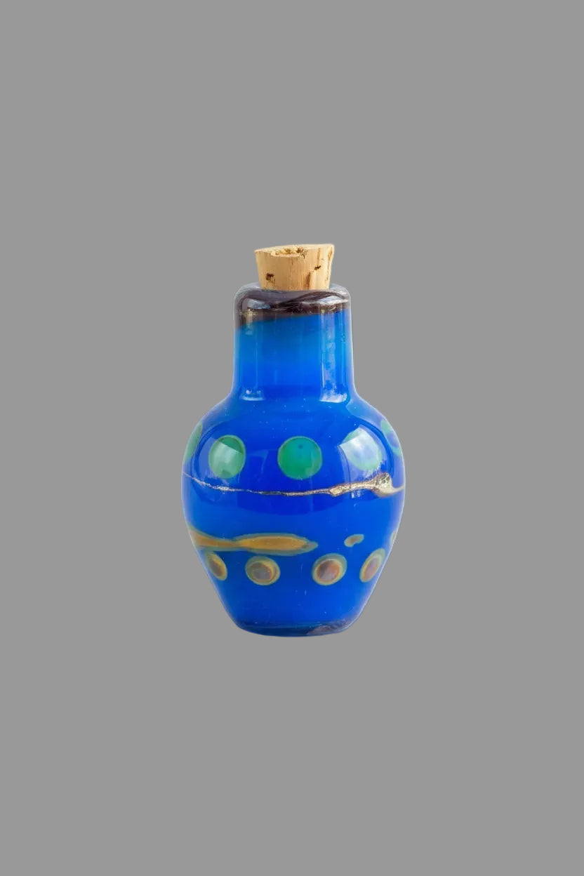A small cobalt blue glass bottle with a cork stopper, decorated with green, gold, and white circular patterns, set against a dark textured background.