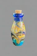 A small glass vial with a cork stopper, featuring a marbled swirl pattern of yellow, blue, black, and iridescent colors, set against a dark textured background.