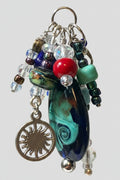 A close-up of a handmade beaded keychain or bag charm hanging from a metal ring. The charm features a large teardrop-shaped glass bead with swirling black, teal, and white patterns, a red bead, clear glass beads, a green bead, and a silver-toned sun charm.