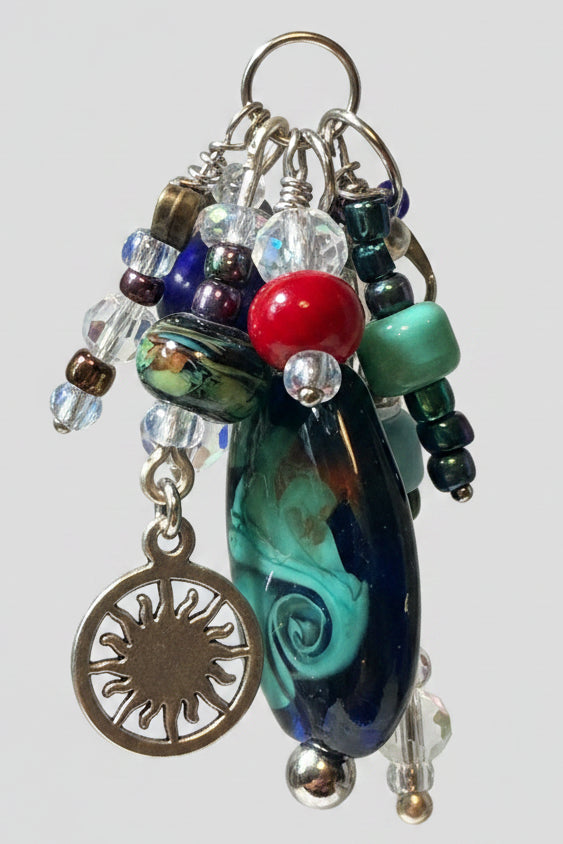 A close-up of a handmade beaded keychain or bag charm hanging from a metal ring. The charm features a large teardrop-shaped glass bead with swirling black, teal, and white patterns, a red bead, clear glass beads, a green bead, and a silver-toned sun charm.