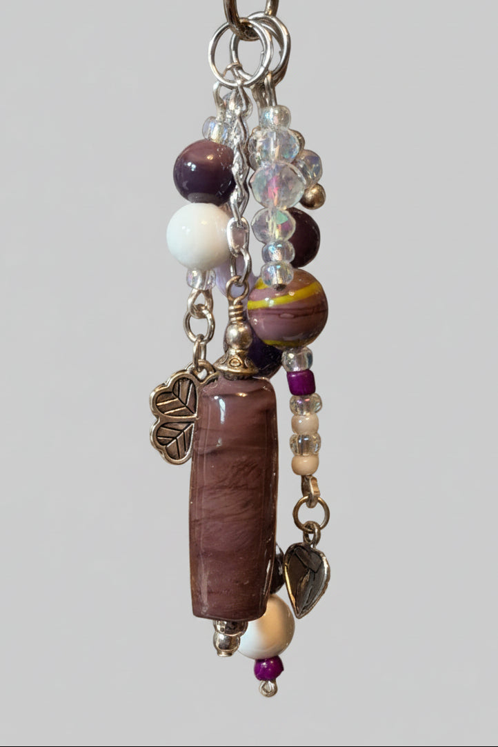 A beaded keychain or bag charm with a long purple stone, white beads, purple beads, clear faceted beads, and silver-toned metal charms.