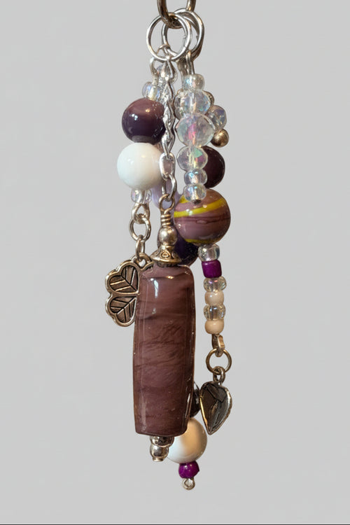 A beaded keychain or bag charm with a long purple stone, white beads, purple beads, clear faceted beads, and silver-toned metal charms.