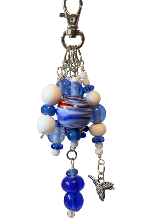 A blue and white beaded bag charm with a marbled glass bead, white beads, blue glass beads, silver metal rings, and a dangling silver bird charm.