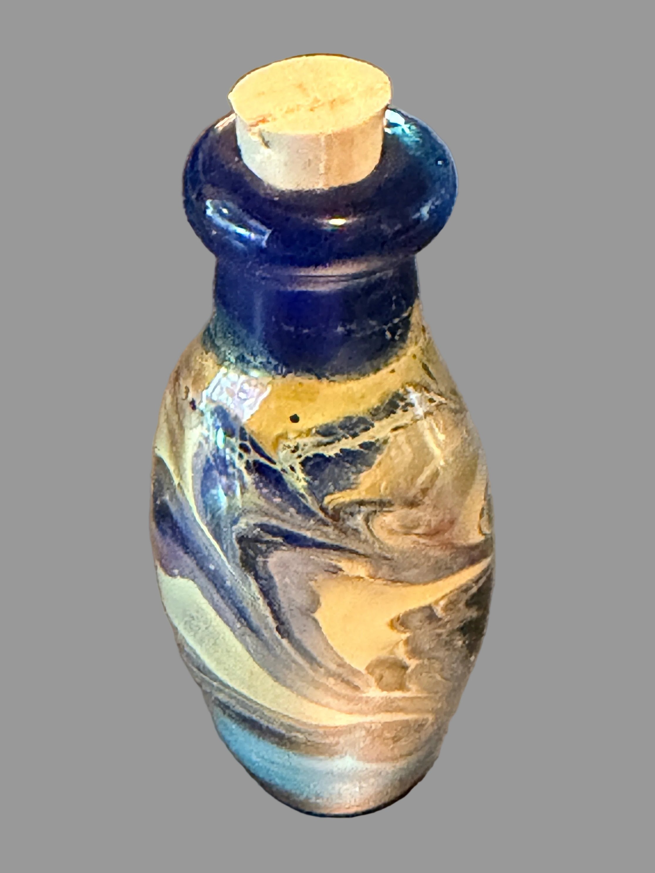 A small decorative glass bottle with a cork stopper, featuring a cobalt blue neck and a marbled body of cream, white, and blue glass.