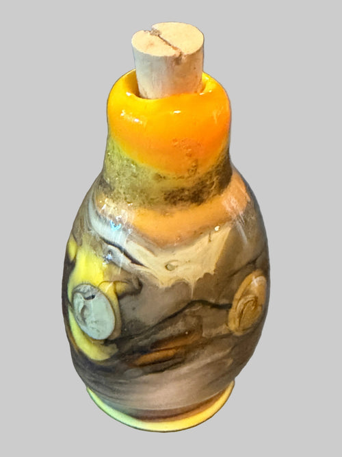 A handcrafted glass bottle with a cork stopper, featuring a marbled pattern of brown, gray, yellow, and white glass with a vibrant orange-yellow section at the neck.
