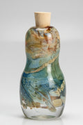 A small handcrafted glass bottle with a cork stopper, featuring a textured, marbled finish in shades of green, blue, and white.