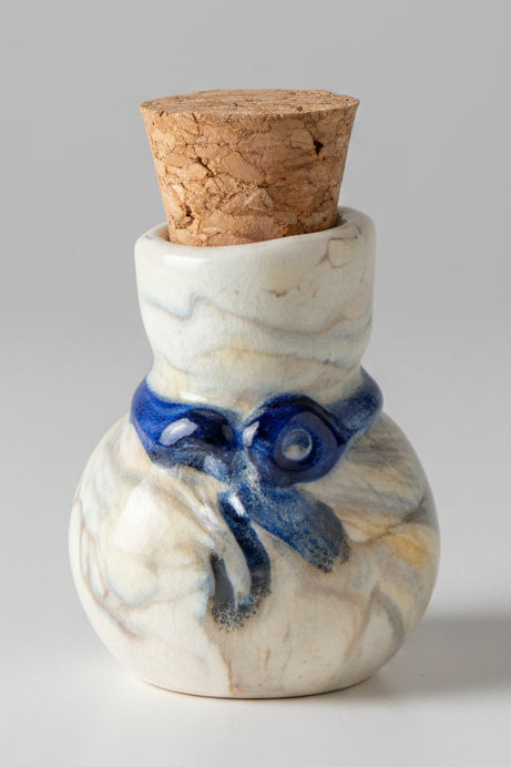 A small glass bottle with a cork stopper, decorated with a blue bow motif on a white marbled glaze, set against a plain gray background.