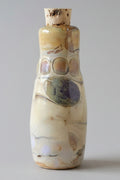 A small handmade ceramic bottle with a cork stopper, featuring a cream and beige body with brown and purple painted accents, set against a plain gray background.