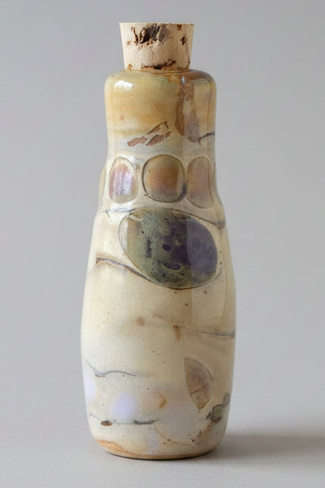 A small handmade ceramic bottle with a cork stopper, featuring a cream and beige body with brown and purple painted accents, set against a plain gray background.