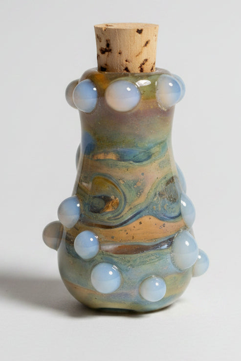A small handmade ceramic bottle with a cork stopper, decorated with a multicolor glaze of blue, green, yellow, and orange, and accented with white bubble-like protrusions, set against a dark gray background.